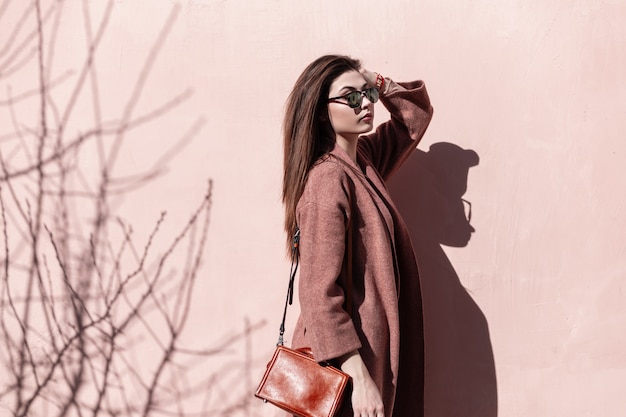 Linda jovem moda modelo elegante em óculos de sol no casaco com bolsa toca o cabelo perto da parede rosa na rua. Linda garota com roupa elegante posa ao ar livre na cidade em um dia ensolarado. Retro.