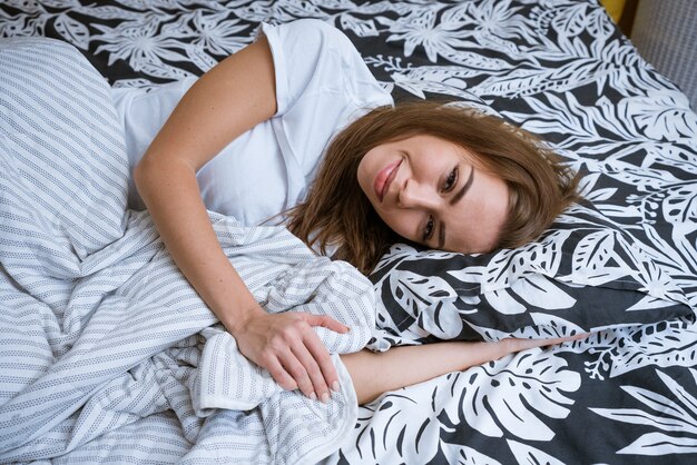 Linda jovem morena acorda de manhã enquanto está deitado em uma cama branca, estilo de vida self-service ...