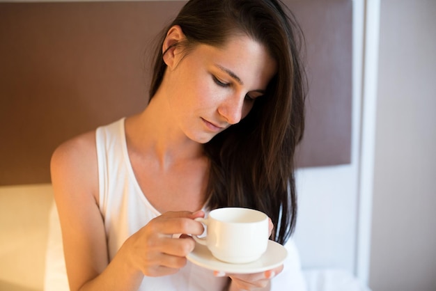 Linda jovem morena bebe café da manhã na cama branca