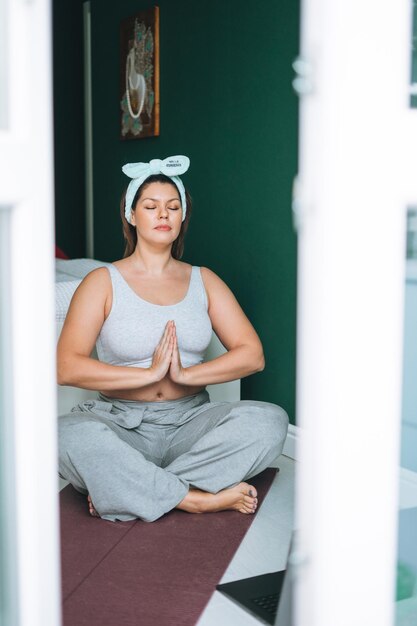 Linda jovem morena com corpo de tamanho positivo em roupas esportivas pratica ioga on-line em casa