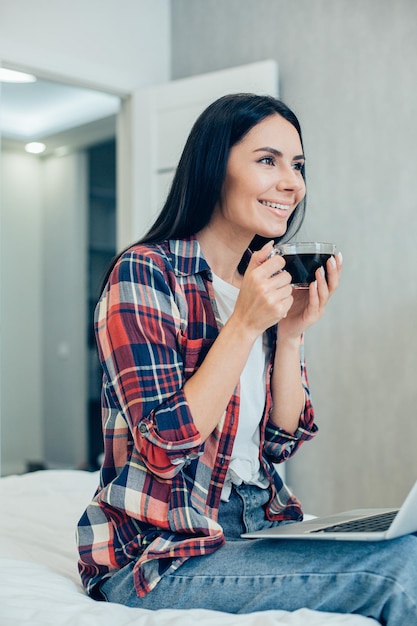 Linda jovem morena com roupas casuais, sentada na cama com uma xícara de café nas mãos e um laptop moderno no colo