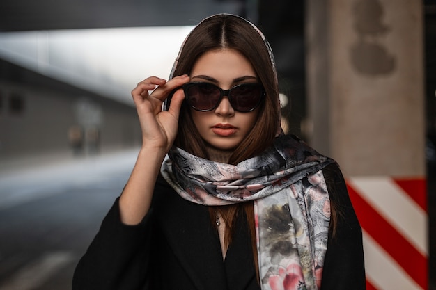 Linda jovem morena com roupas elegantes endireita os óculos de sol. modelo de garota profissional com um elegante casaco preto com um lenço de seda vintage na cabeça, posando na rua perto da estrada.