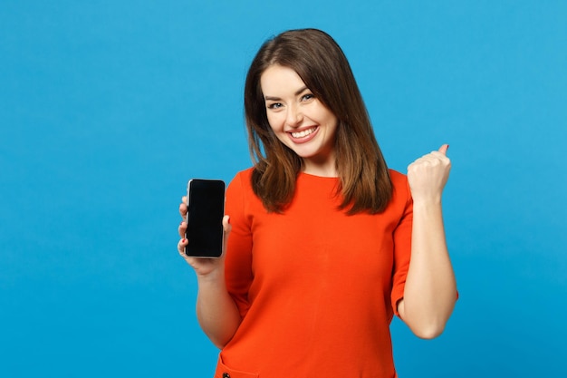 Linda jovem morena de vestido laranja vermelho segura o celular com tela vazia em branco isolada sobre fundo azul na moda, retrato de estúdio. conceito de moda de estilo de vida de pessoas. mock up espaço de cópia.