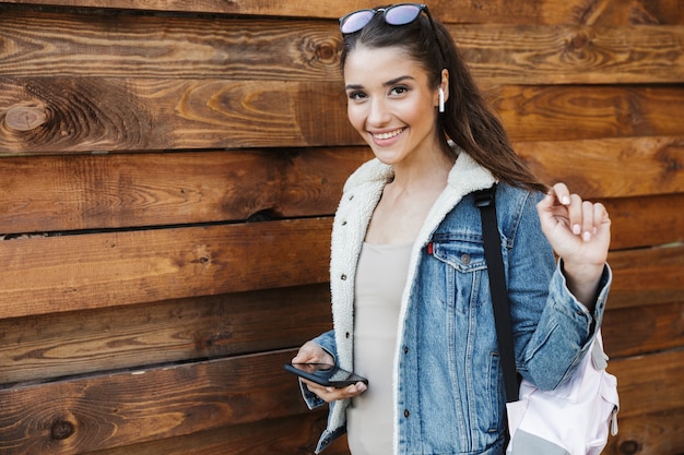 Linda jovem morena vestindo jaqueta, carregando mochila, caminhando ao ar livre pela parede de madeira, ouvindo música com fones de ouvido, segurando o telefone celular