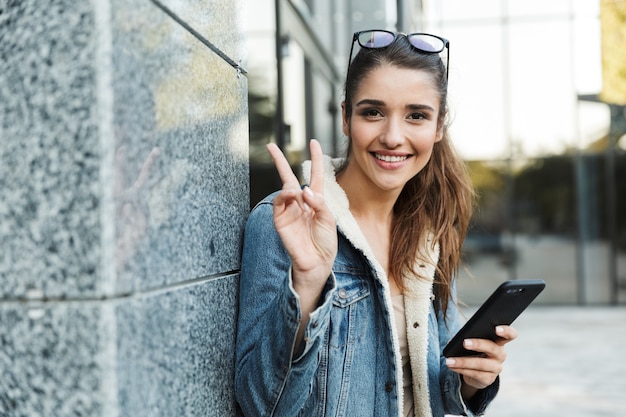 Linda jovem morena vestindo jaqueta, carregando mochila, caminhando ao ar livre, usando telefone celular