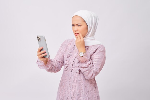 Linda jovem muçulmana asiática assustada usando hijab e vestido roxo lendo más notícias no celular reagindo a más notícias isoladas no fundo branco do estúdio