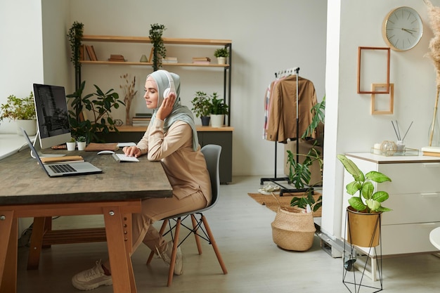 Linda jovem muçulmana usando hijab e fones de ouvido trabalhando remotamente em casa copie o espaço