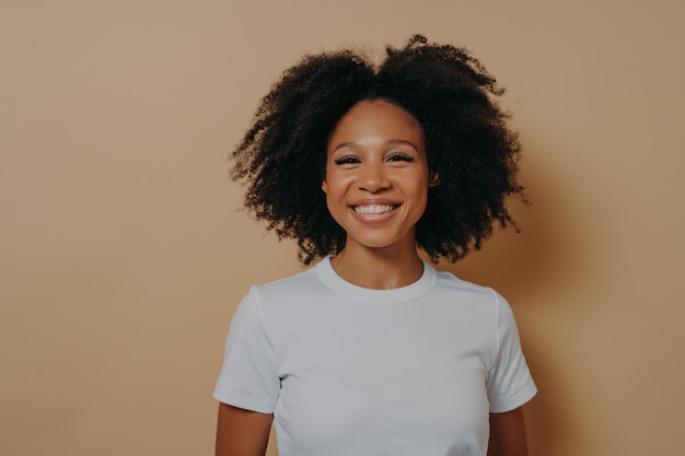 Linda jovem mulher africana sorrindo alegremente para a câmera e demonstrando felicidade, em pé contra a parede colorida no estúdio, mulher morena em roupa casual com cabelos cacheados, mostrando os dentes brancos