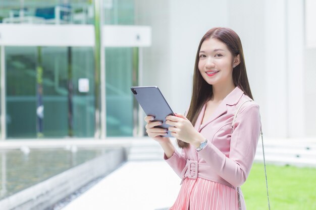 Linda jovem mulher de negócios profissional asiática com cabelo comprido está sorrindo ao ar livre na frente do jardim do escritório enquanto segura o tablet na mão para sair para uma reunião com clientes no escritório.