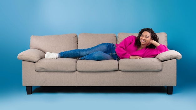 Linda jovem negra feliz em roupas casuais, deitada no sofá e sorrindo para a câmera no estúdio azul