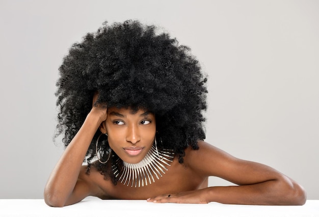 Linda jovem negra modelando uma moderna gargantilha ou colar de prata com brincos de argola, inclinando-se sobre uma mesa com os ombros nus, olhando de lado com um sorriso tranquilo