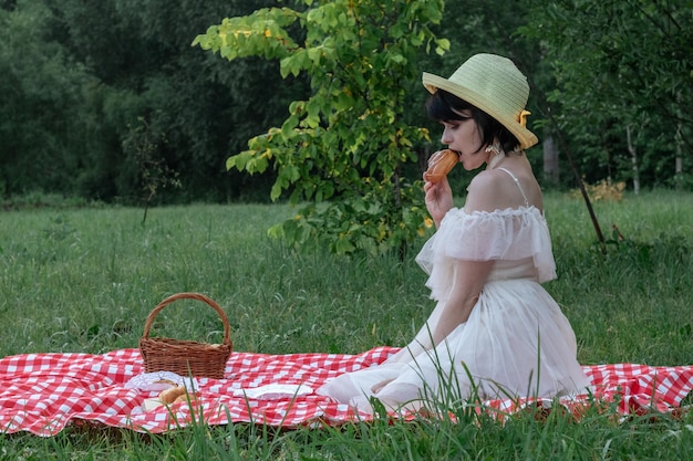 Linda jovem romântica com cabelo curto escuro em um chapéu de palha e vestido branco está sentado em um piquenique na natureza
