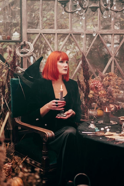 Linda jovem ruiva de chapéu preto e roupa de bruxa faz mágica em estufa decorada para celebração de Halloween com abóboras e crânio na fumaça