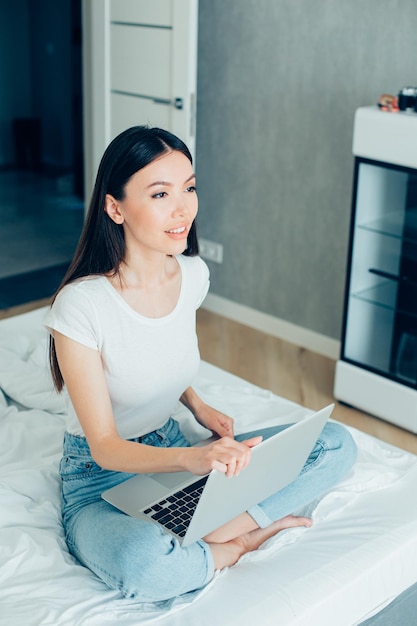 Linda jovem sentada em uma cama de jeans e camiseta e olhando para longe Laptop moderno em seu colo