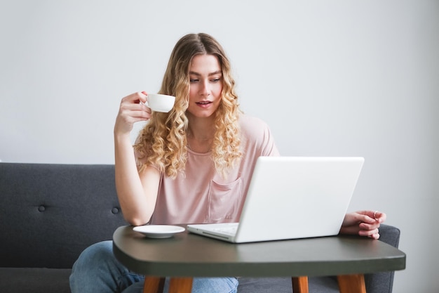 Linda jovem sentada no sofá com uma xícara branca de café trabalhando em um laptop branco Garota em