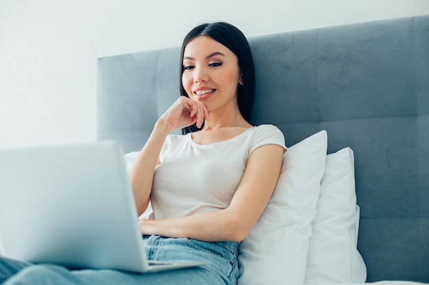 Linda jovem sorridente em uma camiseta branca e jeans sentado em uma cama com um laptop moderno