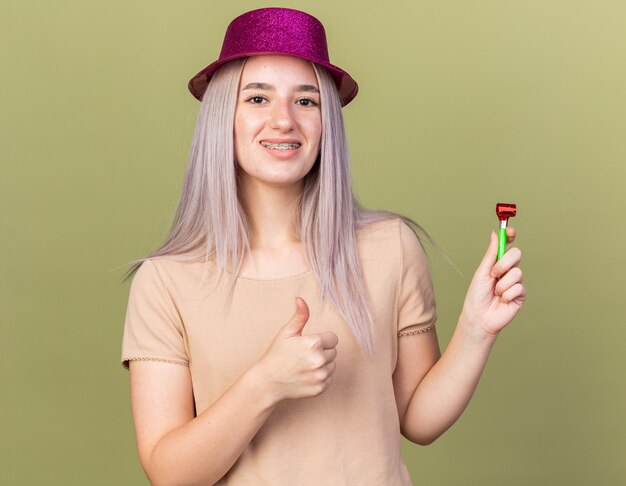 Linda jovem sorridente usando aparelho dentário com chapéu de festa segurando o apito aparecendo o polegar isolado na parede verde oliva