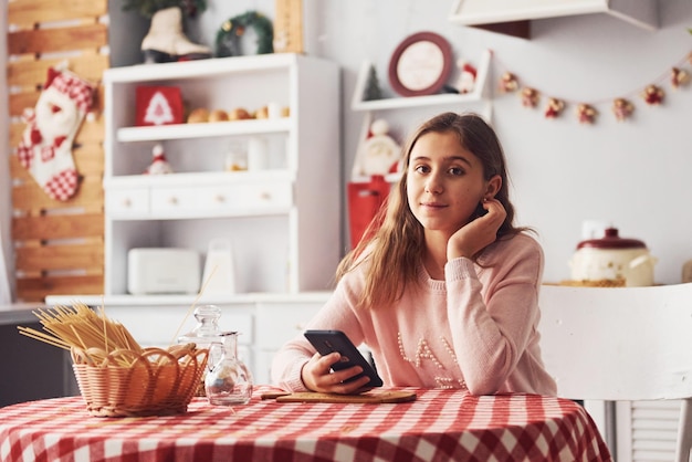 Linda joven adolescente con teléfono en las manos se sienta junto a la mesa en la cocina durante el día.