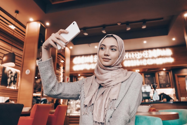 Una linda joven árabe con hiyab se hace selfie en su smartphone en un acogedor restaurante