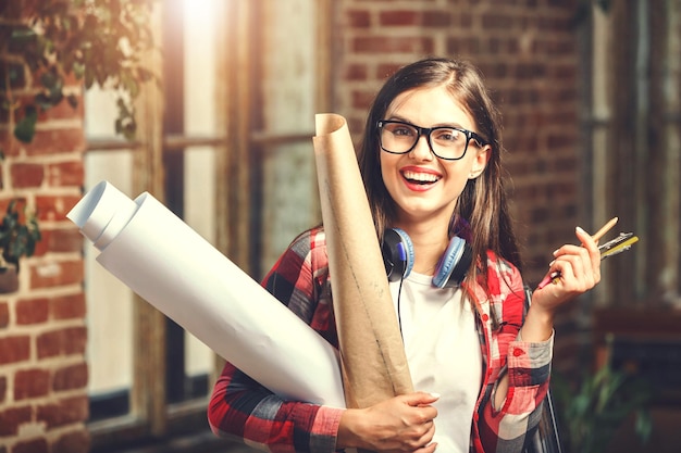 Linda joven arquitecta de pelo largo con gafas con auriculares sosteniendo planos y sonriendo cerca de la ventana en la oficina moderna del loft