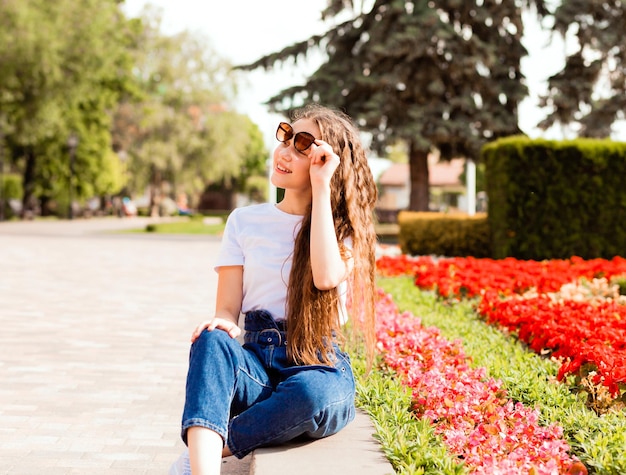 Una linda joven con una camiseta blanca y jeans con gafas de sol se sienta cerca de un lecho de flores