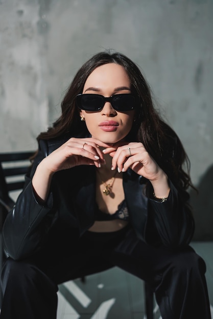 Una linda joven con una chaqueta de cuero y pantalones negros con gafas de sol está sentada en una silla con un fondo gris