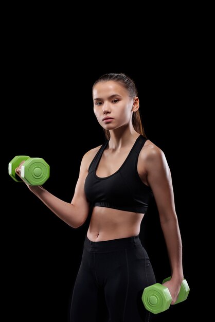 Linda joven deportista en ropa deportiva de pie y haciendo ejercicio con pesas verdes