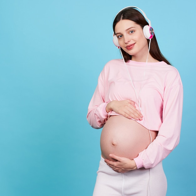 Foto linda joven embarazada escucha música en auriculares sobre un fondo azul.
