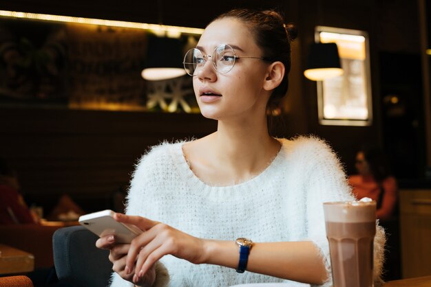 Linda joven estudiante con gafas sentado en la cafetería, bebiendo capuchino después de la escuela, sosteniendo el teléfono inteligente en las manos y mirando por la ventana