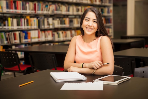 Foto linda joven hispana haciendo algunos deberes en la biblioteca de la escuela y sonriendo