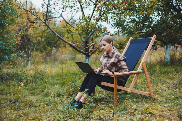 Una linda joven independiente está trabajando en el jardín al aire libre mientras está sentada en una cómoda silla de jardín Trabajo remoto en una computadora portátil