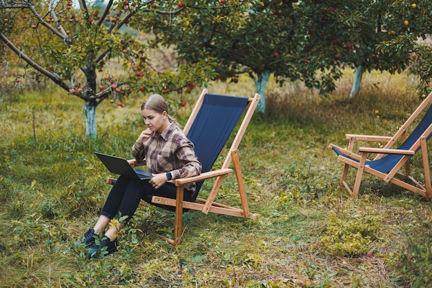 Una linda joven independiente está trabajando en el jardín al aire libre mientras está sentada en una cómoda silla de jardín Trabajo remoto en una computadora portátil