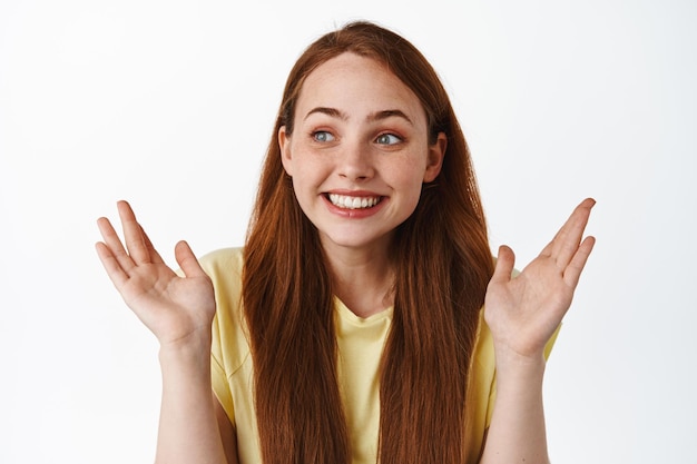 Linda joven mujer moderna con cabello largo rojo, levantando las manos y sonriendo asombrada, mirando a la izquierda en el logo con una mirada impresionada y alegre, fondo blanco