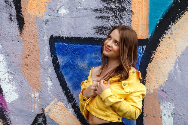 Linda joven mujer sexy de complexión atlética en ropa brillante en la pared decorada. Camina por el centro de la ciudad. Retrato de linda mujer bonita en chaqueta amarilla y pantalón negro en streetwall