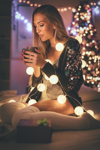 Linda joven mujer sonriente sentada en la cama y sosteniendo una taza de café, rodeada de luces de burbujas navideñas.