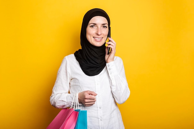 Linda joven musulmana con hiyab sosteniendo bolsas de compras y hablando por teléfono con fondo amarillo Banner