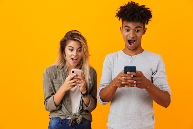 Linda joven pareja amorosa posando aislada sobre pared amarilla mediante teléfonos móviles