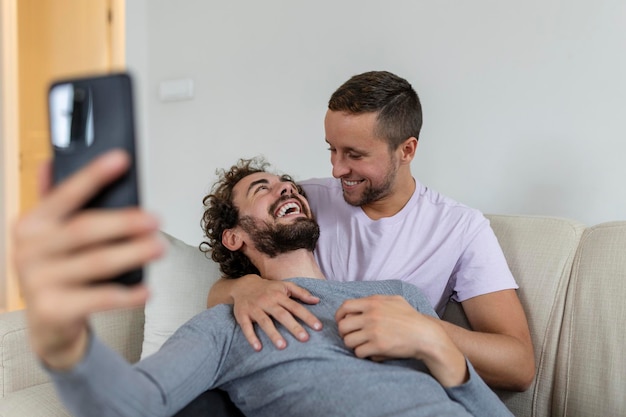 Linda joven pareja gay video llamando a sus amigos en su sala de estar en casa Dos amantes masculinos sonriendo alegremente mientras saludan a sus amigos en un teléfono inteligente Joven pareja gay sentados juntos
