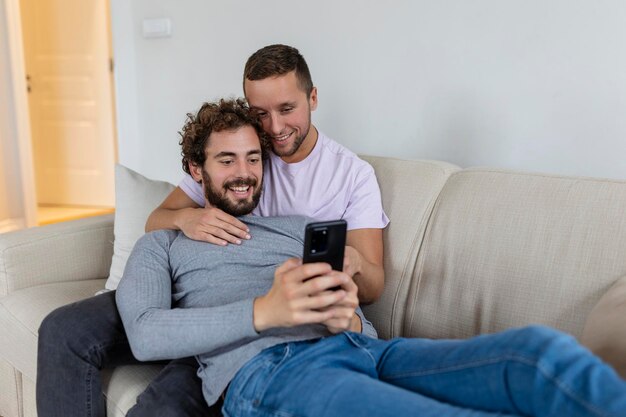 Linda joven pareja gay video llamando a sus amigos en su sala de estar en casa Dos amantes masculinos sonriendo alegremente mientras saludan a sus amigos en un teléfono inteligente Joven pareja gay sentados juntos