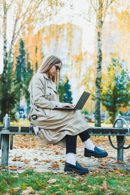 Una linda joven rubia está sentada en un banco en un parque de otoño y trabajando en una computadora portátil Trabajo remoto