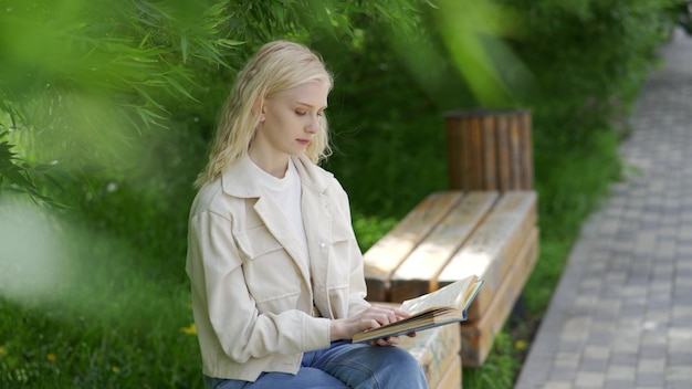 Una linda joven se sienta en un banco del parque y lee un libro sin preocupaciones. Recreación al aire libre. 4K UHD