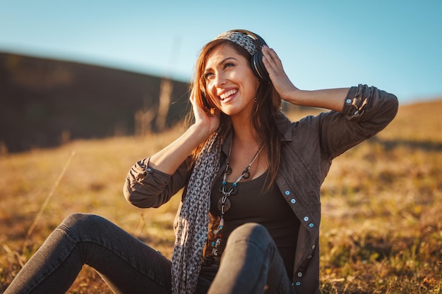 Linda joven sonriente, con auriculares en la cabeza, está sentada en el suelo a principios de primavera y escuchando música.