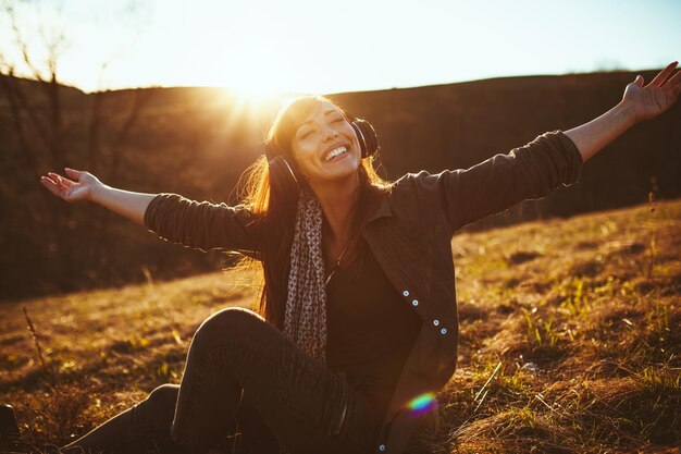 Foto linda joven sonriente, con auriculares en la cabeza, está sentada en el suelo a principios de primavera y escuchando música.