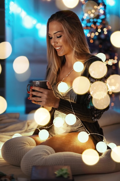 Una linda joven sonriente está sentada en la cama sosteniendo una taza de té, rodeada de luces de burbujas navideñas.