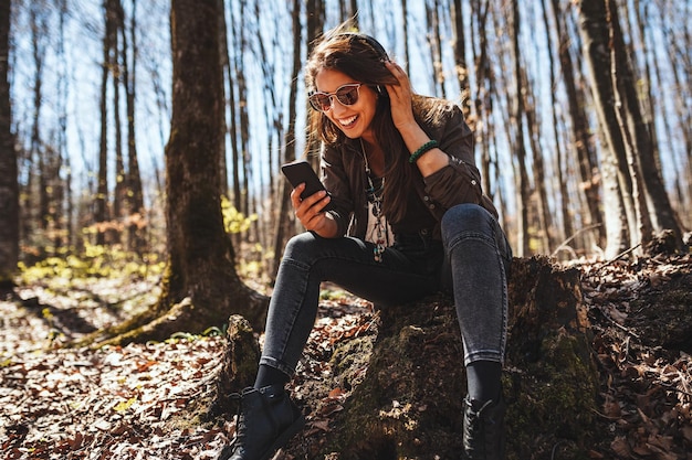 Linda joven sonriente, con un teléfono inteligente en la mano y auriculares en la cabeza, está sentada en las hojas de otoño a principios de otoño y escuchando música.