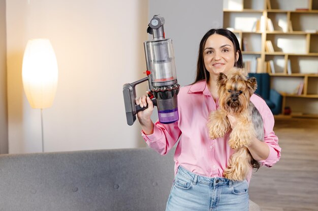 Una linda joven sostiene una aspiradora inalámbrica con un sofá en una mano y un perro en la otra.