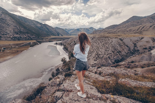 Una linda joven vestida con pantalones cortos de mezclilla y una camisa se para con la espalda y mira el pacífico paisaje montañoso con un arroyo que fluye con las manos dobladas. Admira la vista fascinante.