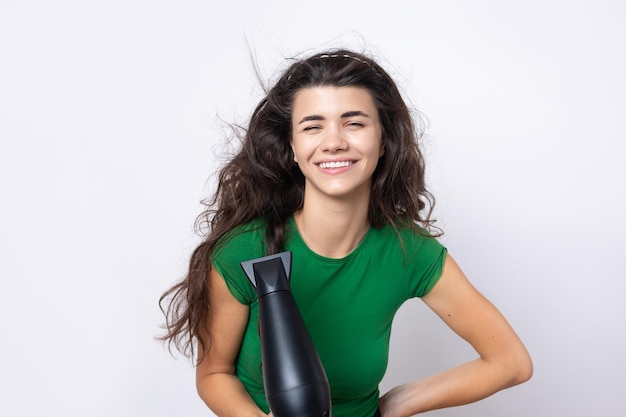 Una linda joven vestida con un top verde seca su hermoso cabello largo y sedoso con un secador de pelo contra un fondo blanco