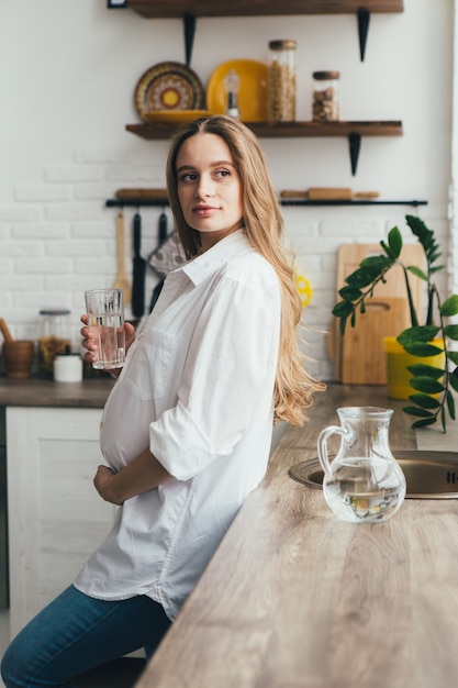 Linda jovencita embarazada bebiendo agua limpia en la cocina