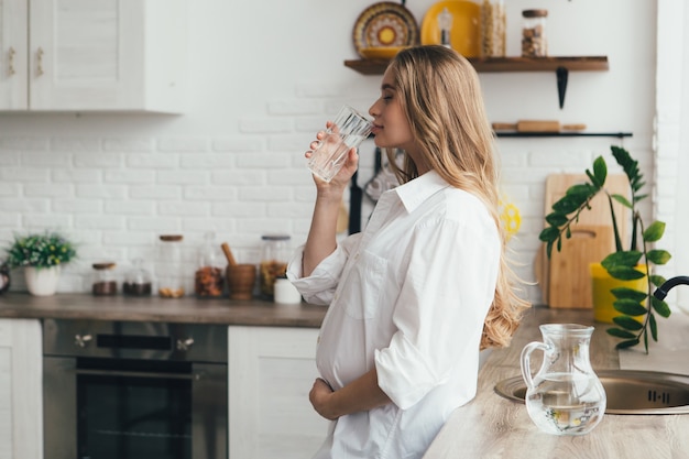 Linda jovencita embarazada bebiendo agua limpia en la cocina